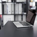 Un ordinateur portable ouvert repose sur un bureau sombre, étagères de classeurs en arrière-plan